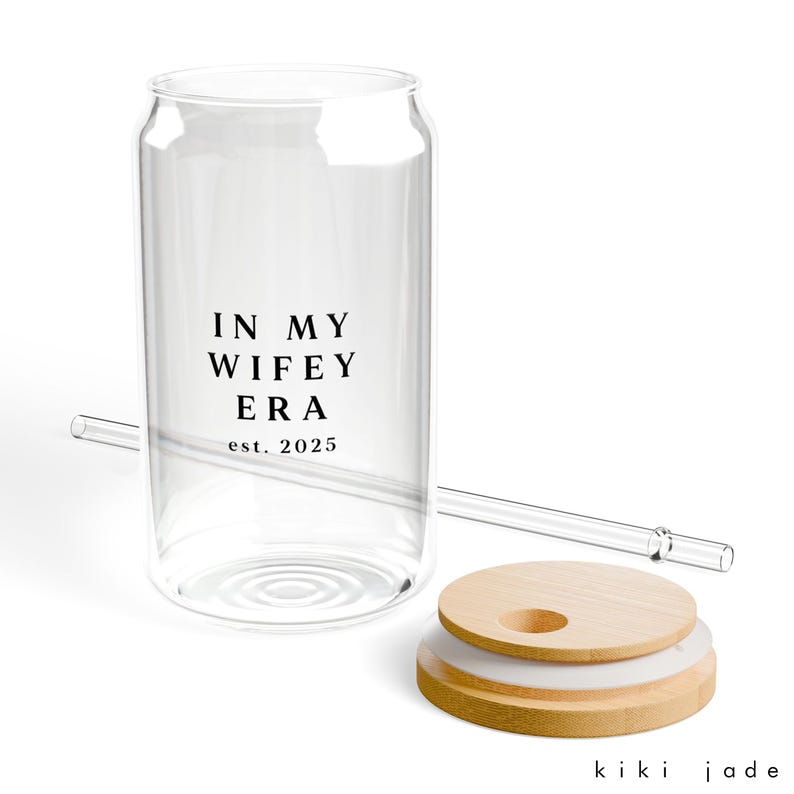 May include: A clear glass can with a black inscription that reads "In My Wife Era est. 2025". The can has a wooden lid with a silicone ring and a clear drinking straw.