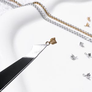 May include: Close-up of jewelry-making tools and components. A gold-colored clasp is held by a silver tool, with a gold and silver rhinestone chain in the background. Several silver clasps are scattered on a white surface.