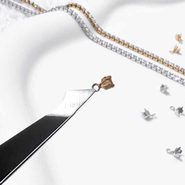 Puede incluir: Primer plano de herramientas y componentes para la fabricación de joyas. Un cierre dorado es sostenido por una herramienta plateada, con una cadena de diamantes de imitación dorada y plateada en el fondo. Varios cierres plateados están dispersos sobre una superficie blanca.