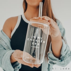 Puede incluir: Una botella de agua de vidrio transparente con tapa de bambú y una pajita. La botella tiene el texto "In My Engaged Era est. 2024" impreso en ella.