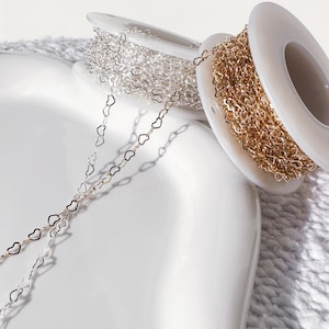 May include: Close-up of spools of heart-shaped chain in silver and gold. The chains are unspooled, with the silver chain extending towards the bottom left. The spools are white, and the background is a soft, neutral color.