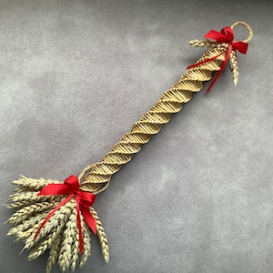 May include: A decorative wheat stalk arrangement. The central section is a tightly woven, spiraling design in a light tan colour. The wheat is tied with red ribbons at both ends, adding a pop of colour. The arrangement is set against a grey background.