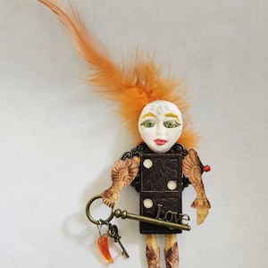 May include: A decorative figurine featuring a white sculpted face with green eyes and red lips, topped with an orange feather. The body is a dark brown domino with the word Love, attached to illustrated arms, legs, and a bronze key charm.