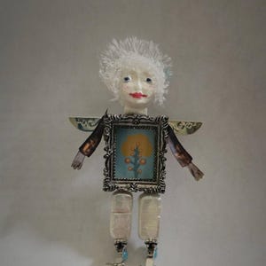 May include: A doll with a white textured head, red lips, and blue eyes. The torso is a rectangular silver frame containing a blue image of a yellow tree. It has dark brown arms, metallic wings, and two white rectangular legs with silver feet.
