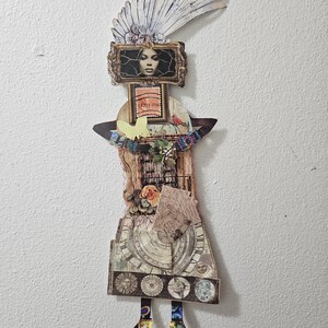 May include: A mixed media wall art piece featuring a collage of a figure with a feathered headdress, a framed portrait of a face, a postage stamp, a yellow butterfly, floral accents, and a skirt decorated with vintage clock and celestial imagery.