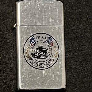 May include: A silver-colored lighter with a brushed metal finish. The lighter features a circular emblem with the text "SSN 713", "Semper Vigilans", and "USS Houston". The emblem includes a submarine illustration and flags.