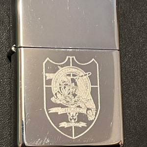 May include: A silver-colored metal lighter with a shield emblem. The emblem features a detailed design with text and a ship. The lighter has a shiny, reflective surface and is closed.