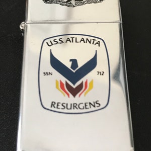 May include: A silver-colored metal lighter with a hinged lid. The lighter features a crest with the text "U.S.S. ATLANTA" and "RESURGENS". The crest includes a blue eagle and other colored accents. A smaller emblem is at the top.