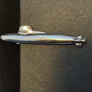May include: A silver-colored submarine tie clip. The clip features a detailed design of a submarine with a conning tower and propellers. The clip is made of a shiny metal and is set against a textured background.