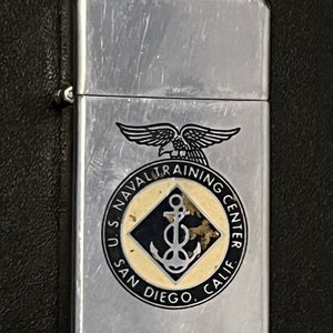 May include: A silver metal lighter with the text "U.S. NAVAL TRAINING CENTER SAN DIEGO, CALIF." and an emblem featuring an eagle and anchor. The lighter has a rectangular shape and a hinged lid.