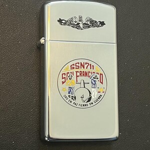 May include: A silver-colored lighter with a detailed emblem featuring the text "SSN711 SAN FRANCISCO" and a submarine illustration. The top of the lighter has a black and white emblem. The lighter has the phrase "ORO EN PAZ FIERRO EN GUERRA".