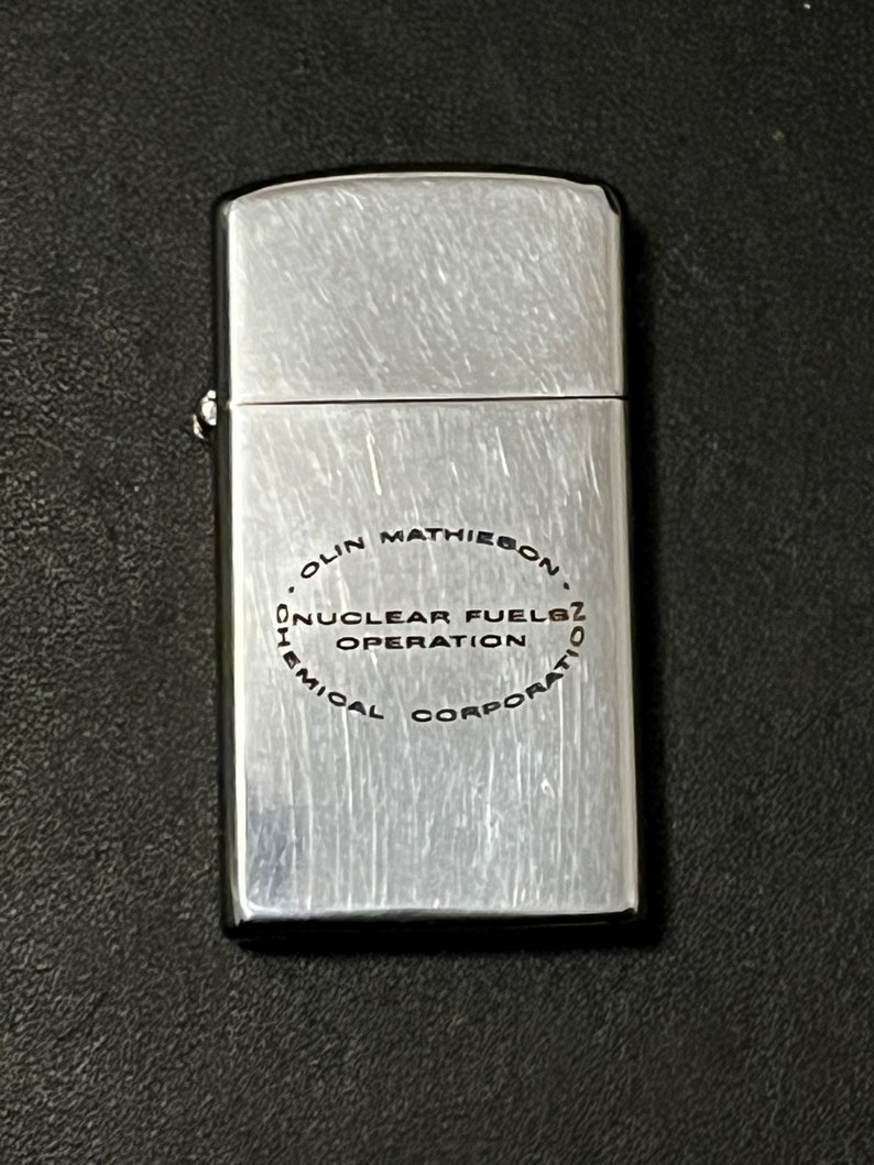 Może przedstawiać: Srebrna zapalniczka z wykończeniem ze szczotkowanego metalu. Zapalniczka ma napis "OLIN MATHIESON NUCLEAR FUELS OPERATION CHEMICAL CORPORATION" wydrukowany w okręgu.