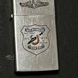 May include: A silver-colored lighter with a detailed emblem featuring the text "USS PLUNGER" and "SSM 595". The lighter has a hinged lid and a textured surface. The emblem includes a horseshoe, arrow, and the text "WHAT IS FAST" and "IS PROLOGUE".