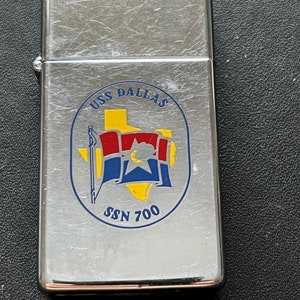 May include: A silver-colored lighter with the text "USS DALLAS" and a circular emblem featuring a Texas-shaped yellow background, red and blue flags, and the text "SSN 700". The lighter has a brushed metal finish.