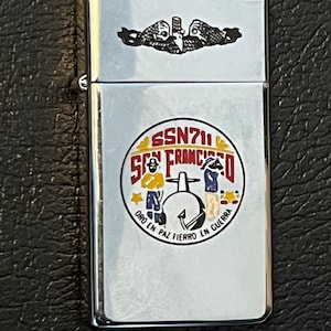 May include: A silver-colored lighter with a detailed emblem featuring the text "SSN711 SAN FRANCISCO" in a circular design. The lighter also has a black and white emblem at the top. The design includes figures and the phrase "ORO EN PAZ FIERRO EN GUERRA".