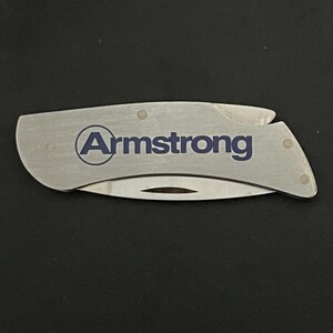 May include: A silver folding pocket knife with the word "Armstrong" printed in blue. The knife is closed and has a curved shape with a blade and a handle. The background is black.