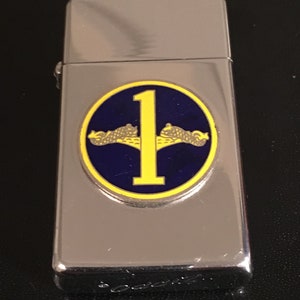 May include: A silver-colored lighter with a circular emblem featuring a yellow number "1" and two yellow submarines on a blue background. The lighter has a shiny, reflective surface and a hinged lid.