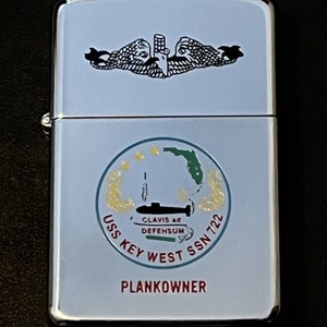 May include: A silver lighter with a light blue finish. The lighter features a black emblem of two stylized fish and a submarine. The text "USS KEY WEST SSN 722" and "PLANKOWNER" are also visible.