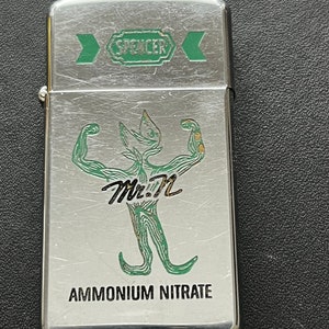 Pode incluir: Um isqueiro prateado com um gráfico verde e preto de uma figura musculosa com o texto "Mr. N" e "Ammonium Nitrate". A parte superior do isqueiro tem o texto "Spencer" num design verde.