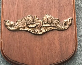 Puede incluir: Placa de madera con forma de escudo. Un emblema metálico dorado de dos delfines que flanquean un objeto central está fijado a la placa. La madera tiene un tono marrón medio.