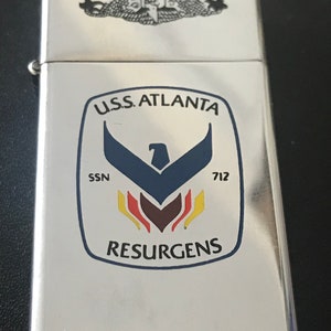 May include: A silver metal lighter with a rectangular shape. The lighter features a crest with the text "U.S.S. ATLANTA" and "RESURGENS". The crest includes a blue eagle design and the number "712". The top of the lighter has a decorative emblem.