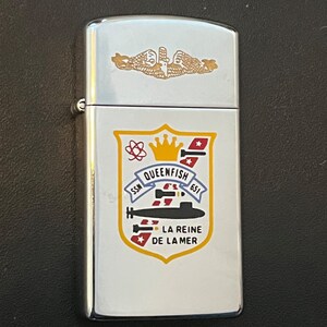 May include: A silver-colored lighter with a gold emblem on the top. The front features a crest with the text "SSN QUEENFISH 651" and "LA REINE DE LA MER". The crest includes a submarine illustration, a crown, and red and yellow accents.