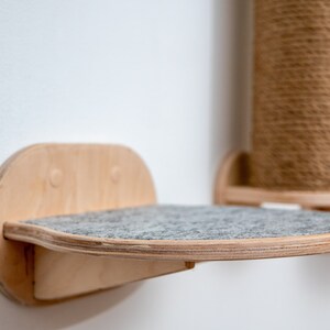 Puede incluir: Una estanter&iacute;a para gatos de madera con una superficie de fieltro gris. La estanter&iacute;a est&aacute; montada en una pared blanca y tiene una forma redondeada.