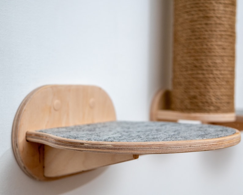 Puede incluir: Una estanter&iacute;a para gatos de madera con una superficie de fieltro gris. La estanter&iacute;a est&aacute; montada en una pared blanca y tiene una forma redondeada.