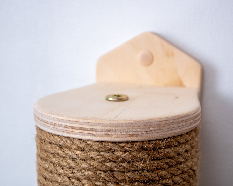 Puede incluir: Un rascador para gatos de madera con una base cubierta de cuerda y una plataforma de madera en la parte superior. La plataforma tiene un tornillo en el centro.