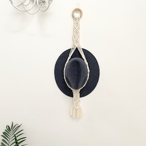 May include: A navy blue hat held by a cream macrame hat hanger. The hanger has a wooden ring at the top and detailed knotting. The hat is round with a curved brim. The background is a plain white wall.