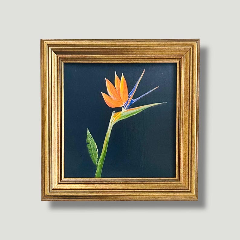 May include: A framed painting of a bird of paradise flower. The flower features orange petals, blue accents, and a green stem and leaves. The painting is set against a dark blue background and framed in a gold-coloured frame.
