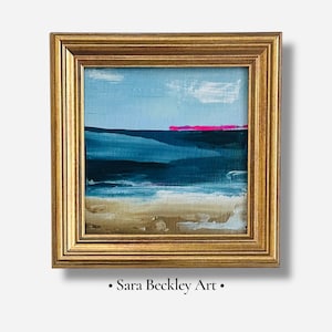 May include: Abstract landscape painting in a gold frame. The artwork features a blue sky with a few white clouds, a dark blue sea, and a pink horizon line. The bottom of the painting depicts a sandy beach. The text "Sara Beckley Art" is at the bottom.