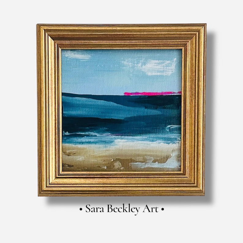 May include: Abstract landscape painting in a gold frame. The artwork features a blue sky with a few white clouds, a dark blue sea, and a pink horizon line. The bottom of the painting depicts a sandy beach. The text "Sara Beckley Art" is at the bottom.