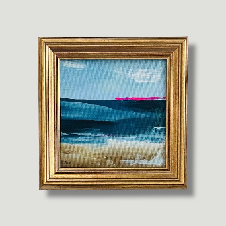 May include: Small framed abstract landscape painting. The artwork features a blue sky with white clouds, a dark blue ocean, and a tan beach. A bright pink line accents the horizon. The painting is in a gold frame.