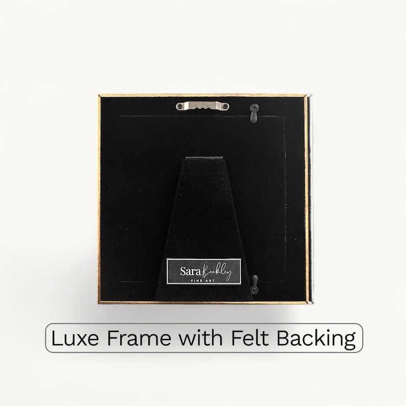 K&ouml;nnte beinhalten: Quadratischer Bilderrahmen mit goldfarbenem Rand und schwarzer R&uuml;ckseite. Der Rahmen hat eine Filzr&uuml;ckseite und einen kleinen Metallaufh&auml;nger. Der Text "Luxe Frame with Felt Backing" befindet sich unten.