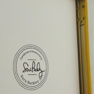 K&ouml;nnte beinhalten: Ein gerahmtes Kunstwerk mit einem goldfarbenen Metallrahmen. Das Kunstwerk hat einen wei&szlig;en Hintergrund mit einem kreisf&ouml;rmigen schwarzen Stempel mit der Aufschrift "Certificate of Authenticity" und der Signatur des K&uuml;nstlers "Sara Beckley Art."