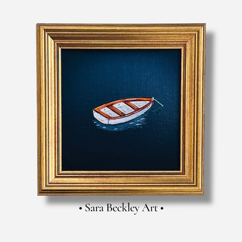 May include: A framed painting of a small white and orange boat on dark blue water. The painting is in a gold frame. The text "Sara Beckley Art" is at the bottom of the frame.