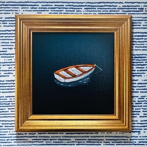 May include: A framed painting depicting a small white and orange boat on dark blue water. The boat is secured with a green rope. The painting is in a gold-coloured frame, set against a blue and white striped background.