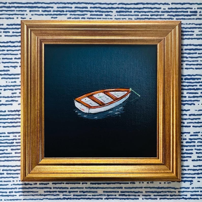 May include: A framed painting depicting a small white and orange boat on dark blue water. The boat is secured with a green rope. The painting is in a gold-coloured frame, set against a blue and white striped background.