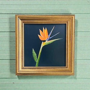 May include: A framed painting of a Bird of Paradise flower. The flower has orange, blue, and green hues against a dark blue background. The painting is in a gold-coloured frame and is mounted on a light green wooden surface.