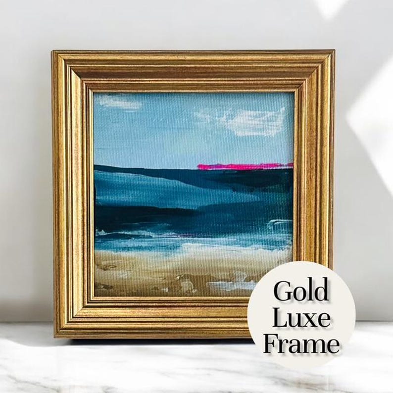 May include: A framed abstract landscape painting with a gold-colored frame. The artwork features a blue sky with white clouds, a dark blue body of water, and a strip of pink. The text "Gold Luxe Frame" is on a white circle.