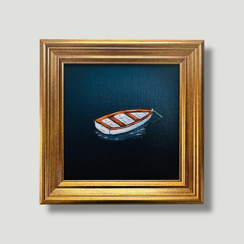 May include: A small painting of a white and orange rowboat on dark blue water, framed in a gold-coloured, ornate frame. The boat is tied to a green rope. The painting is square and set against a white background. The artwork is a nautical theme.