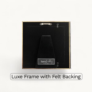 May include: Square picture frame with a gold border and black backing. The frame has a felt backing and a small metal hanger. The text "Luxe Frame with Felt Backing" is at the bottom.