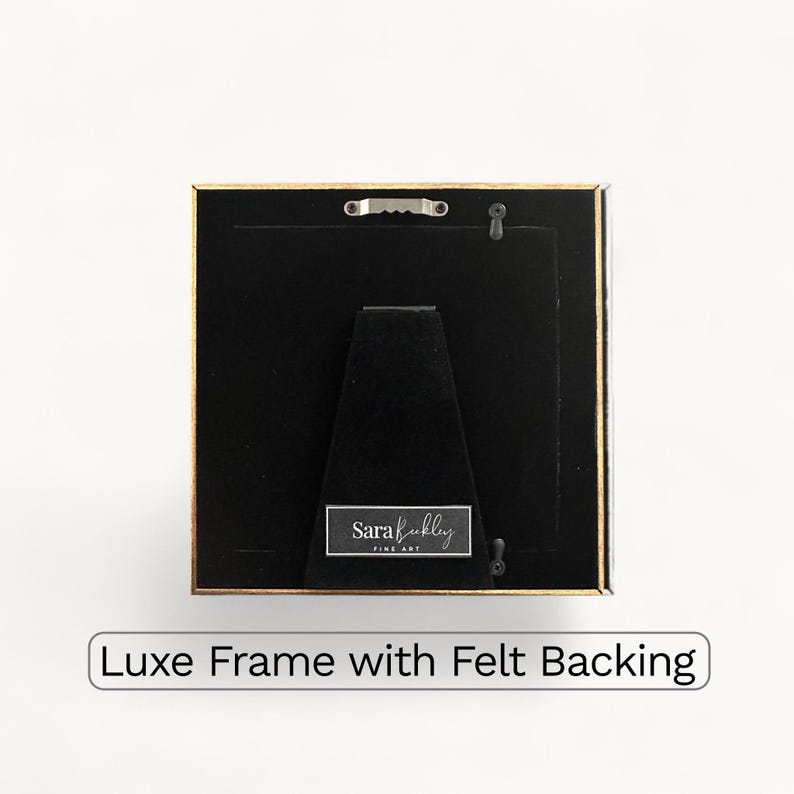 May include: Square picture frame with a gold border and black backing. The frame has a felt backing and a small metal hanger. The text "Luxe Frame with Felt Backing" is at the bottom.