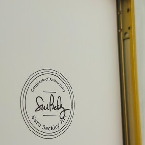 May include: A framed artwork with a gold-coloured metal frame. The artwork has a white background with a circular black stamp that reads "Certificate of Authenticity" and the artist's signature, "Sara Beckley Art."