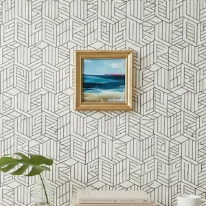May include: A framed painting of a seascape with blue and turquoise water and a sandy beach hangs on a wall with a black and white geometric pattern. A monstera leaf in a glass vase and a stack of books are on a wooden surface.