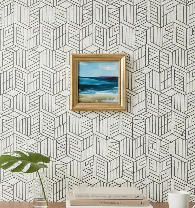May include: A framed painting of a seascape with blue and turquoise water and a sandy beach hangs on a wall with a black and white geometric pattern. A monstera leaf in a glass vase and a stack of books are on a wooden surface.