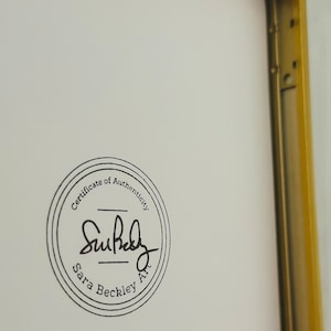 May include: A framed artwork with a gold-coloured metal frame. The artwork has a white background with a circular black stamp that reads "Certificate of Authenticity" and the artist's signature, "Sara Beckley Art."