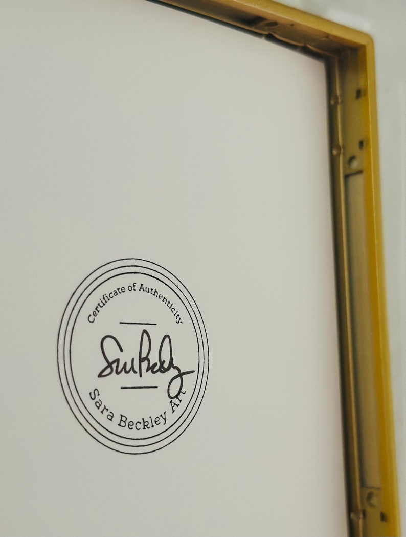 May include: A framed artwork with a gold-coloured metal frame. The artwork has a white background with a circular black stamp that reads "Certificate of Authenticity" and the artist's signature, "Sara Beckley Art."