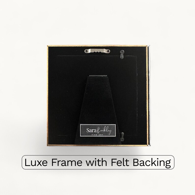 May include: Square picture frame with a gold-colored border and black backing. The frame has a felt backing and a small metal hanger. The text "Luxe Frame with Felt Backing" is at the bottom.